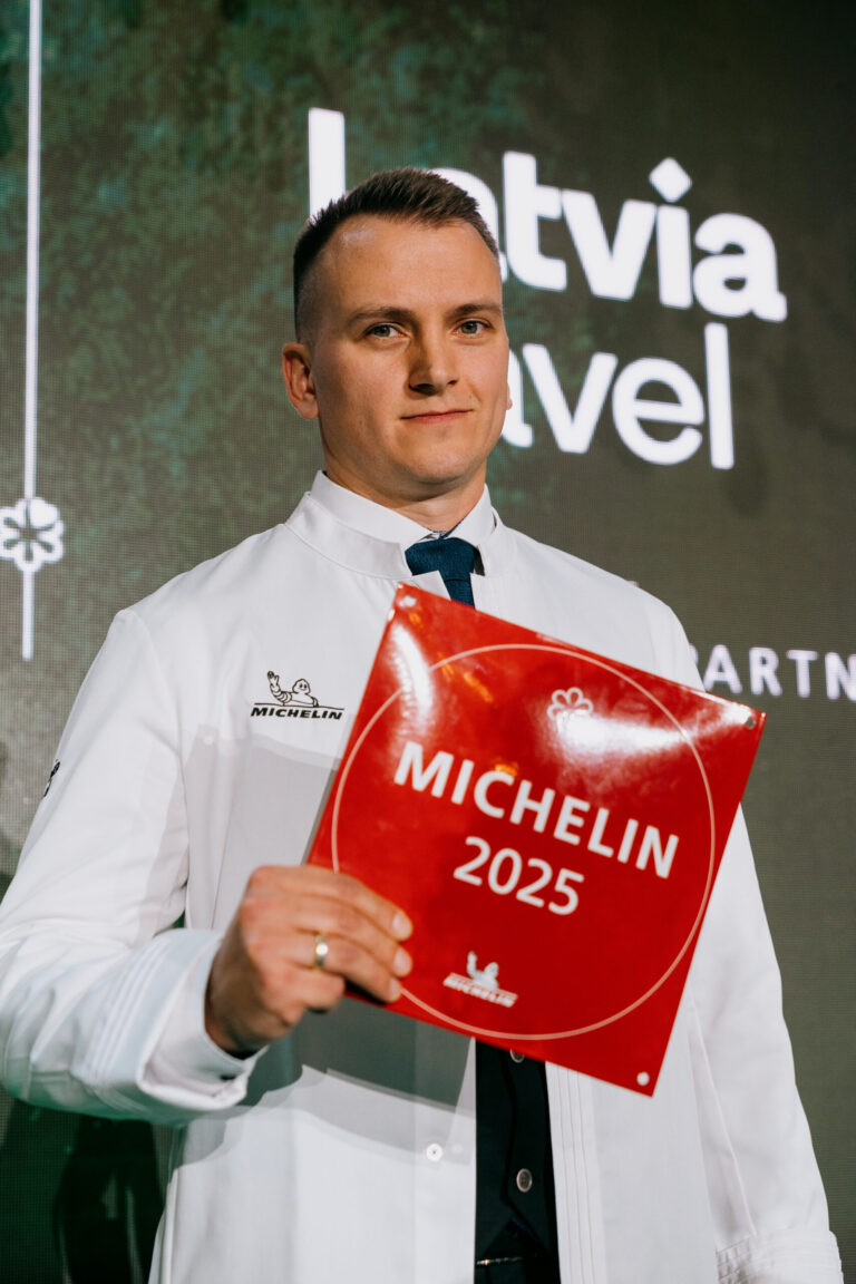 Kristaps Silis hoiab käes punast MICHELIN 2025 plaati, kandes valget kokarõivast, millel on Michelini logo, sümboliseerides kulinaarse tipptaseme tunnustust.